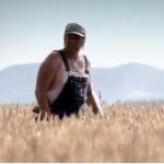 man standing in field wearing overalls, wifebeater tshirt, hat